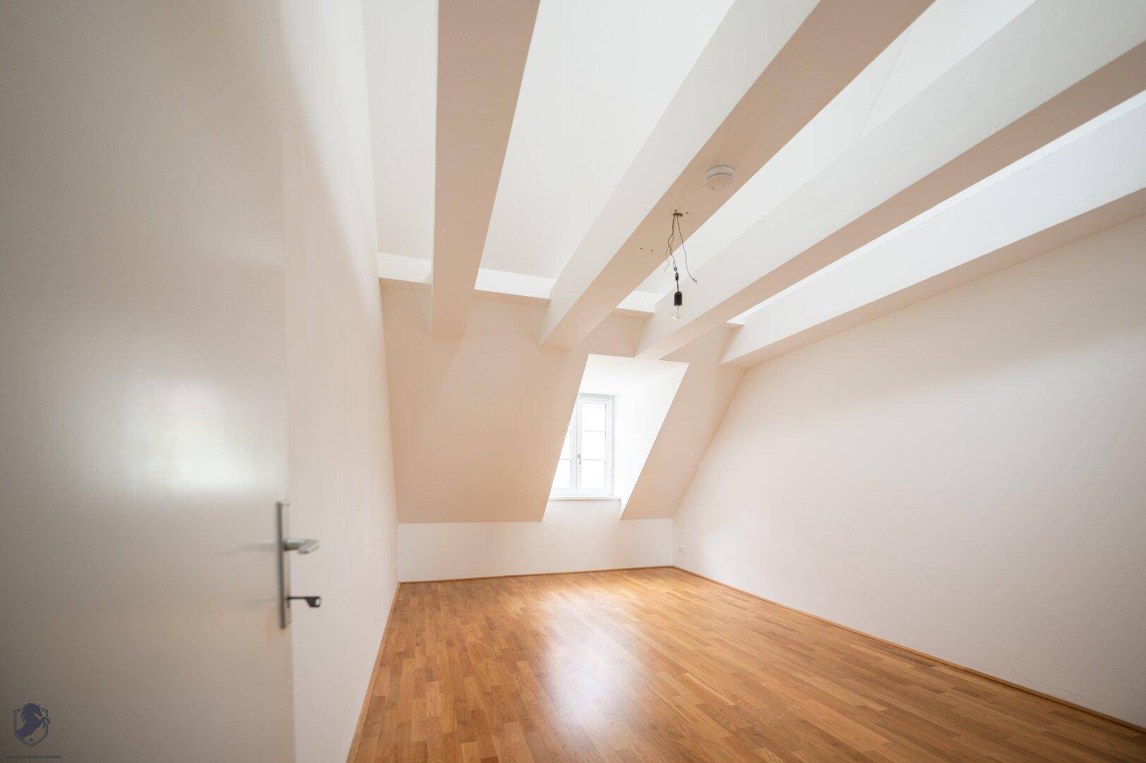 Geräumiger Raum mit Holzboden, weißen Wänden und schrägen Decken im Dachgeschoss.