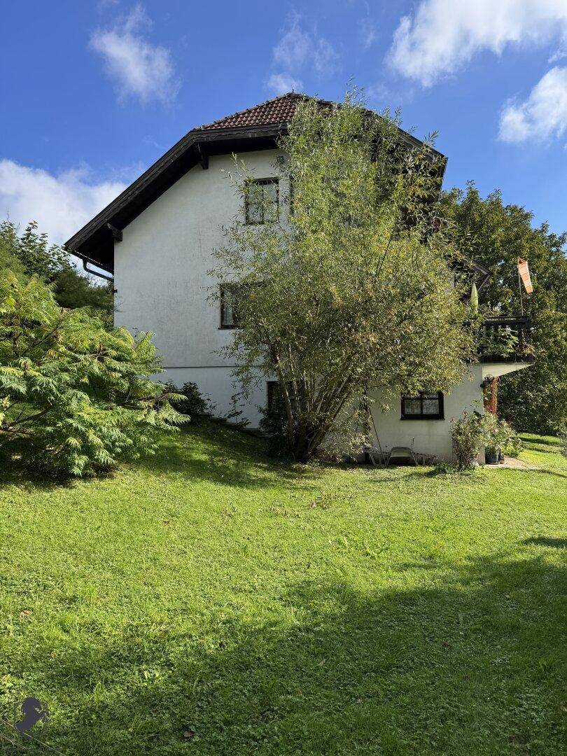 Hausfassade mit Fenstern und Balkon, umgeben von einem großen, grünen Garten.