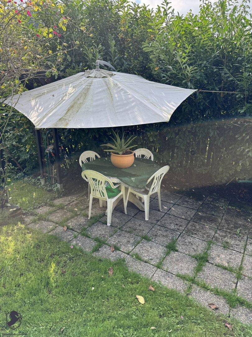 Einfache Terrasse mit Gartentisch, Stühlen und Sonnenschirm, umgeben von Hecken.