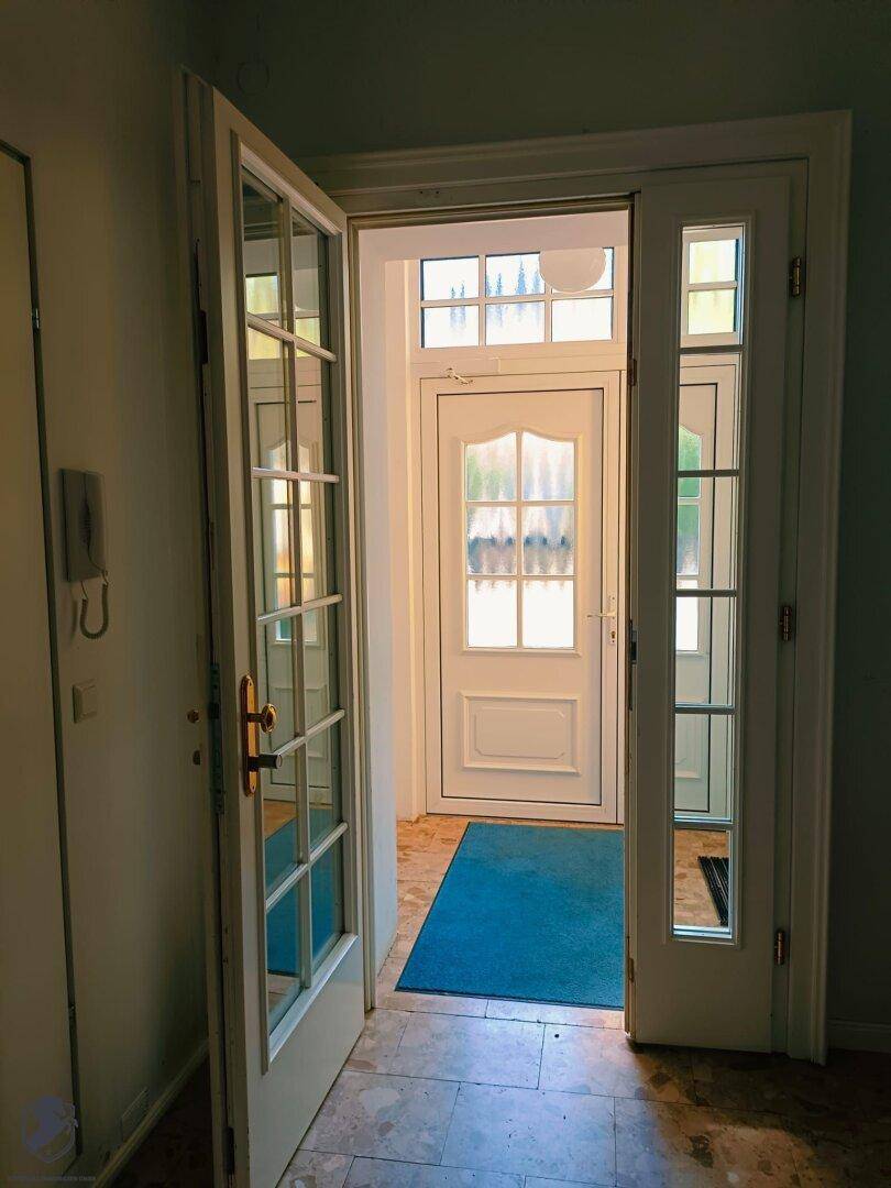 Heller Eingangsbereich mit einer Doppeltür und einem blauen Teppich, führt ins Innere.