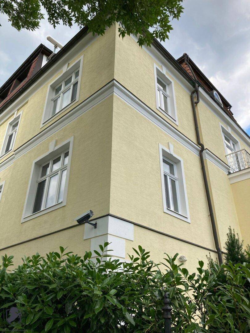 Ecke der gelben Villa mit Überwachungskamera und grünen Hecken im Vordergrund.