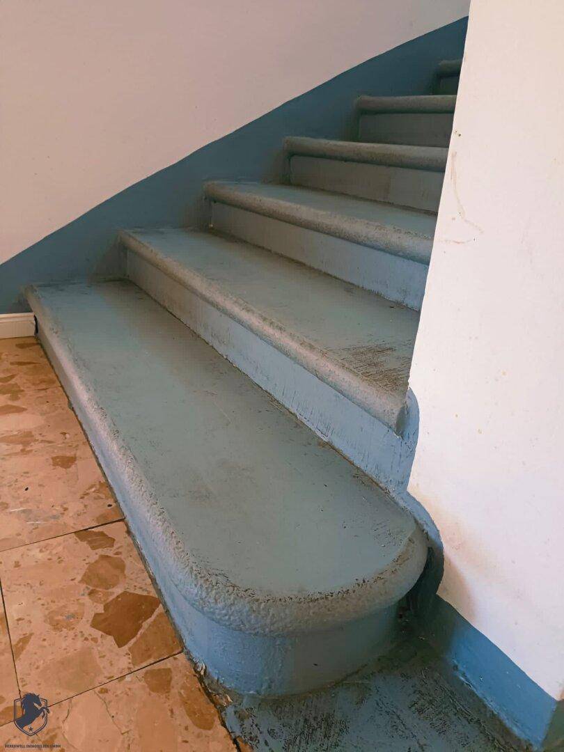 Treppenaufgang mit blauen Stufen und Terrazzoboden, führt in die oberen Etagen.