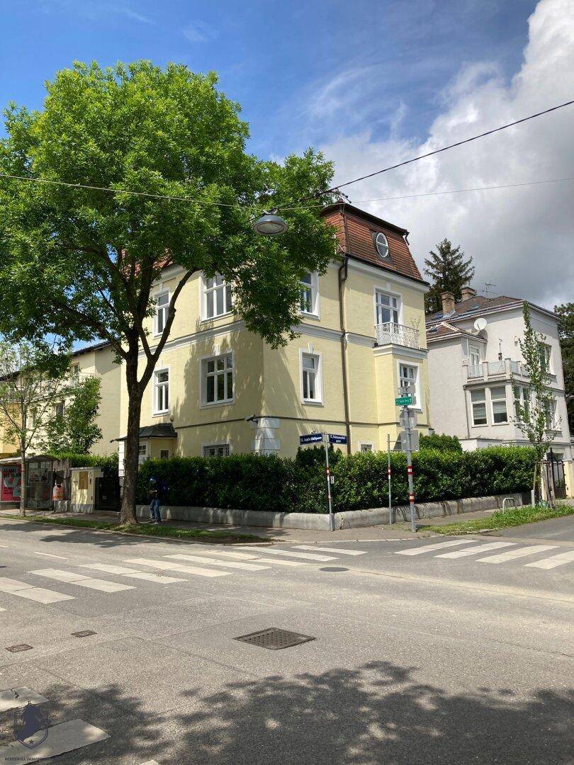 Die gelbe Villa an einer Straßenecke, umgeben von Bäumen und Hecken, unter einem sonnigen Himmel.