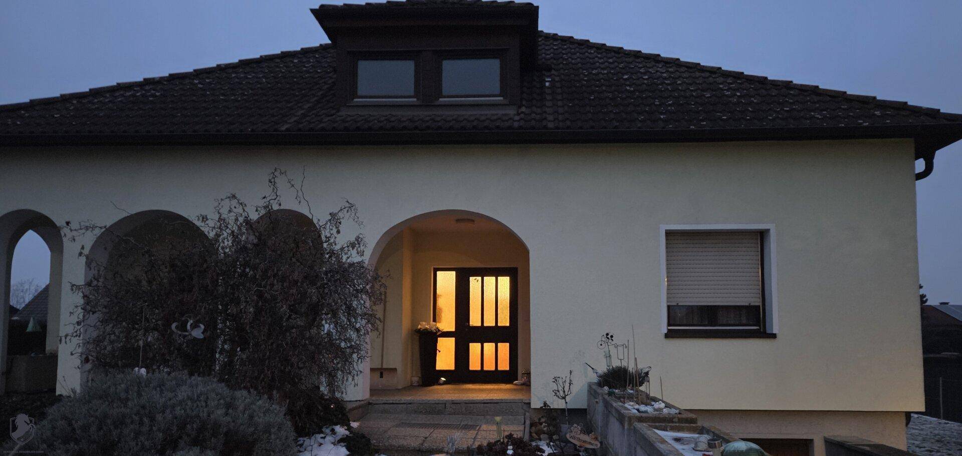 Einfamilienhaus bei Dämmerung mit beleuchtetem Eingangsbereich, Dachgaube und Rollläden.