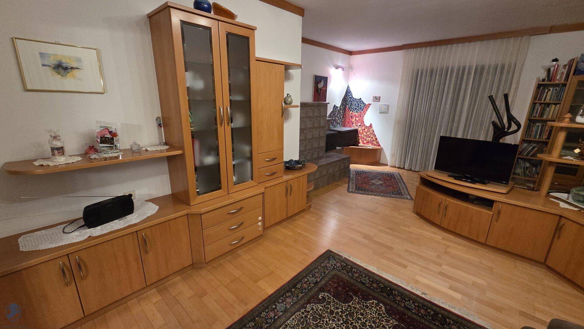 Geräumiges Wohnzimmer mit Parkettboden, Holzschränken und einem traditionellen Kachelofen.