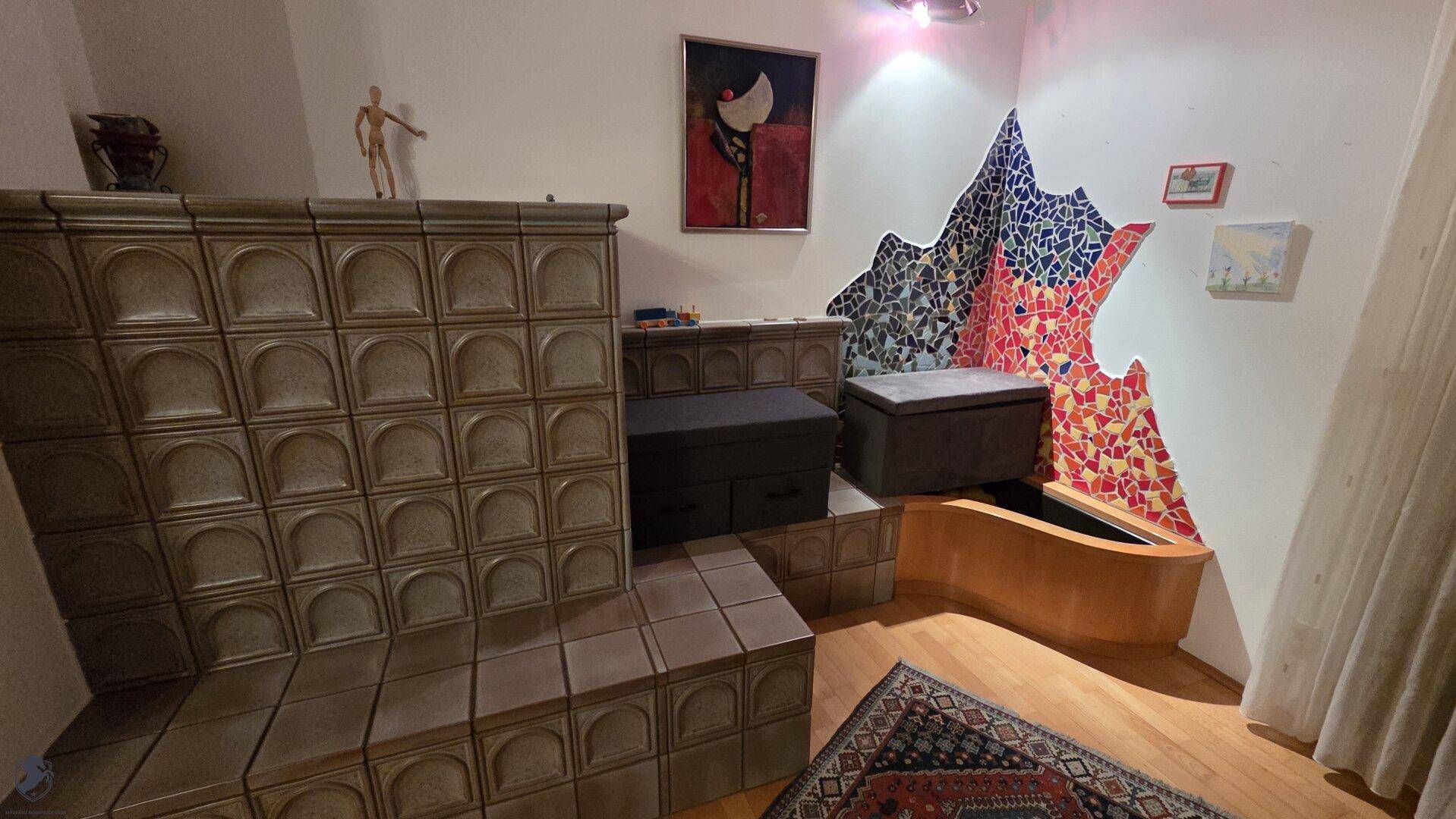 Detailansicht des Kachelofens im Wohnzimmer mit farbenfroher Wanddekoration und Holzboden.