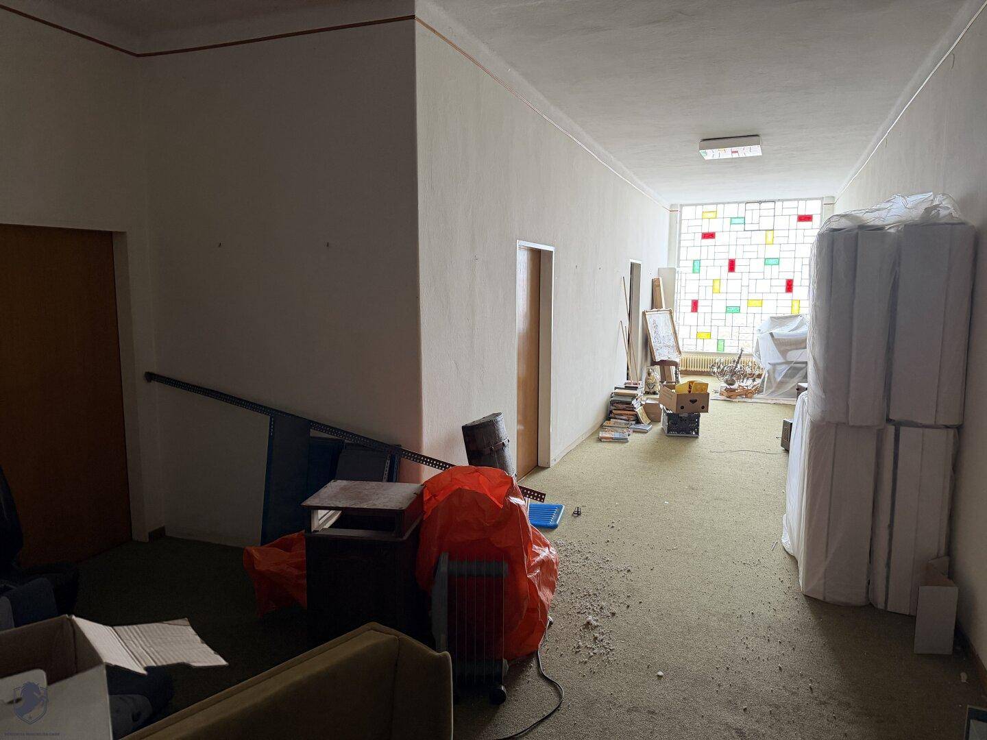 Langer, schmaler Flur mit Teppichboden und einem farbenfrohen Buntglasfenster am Ende.