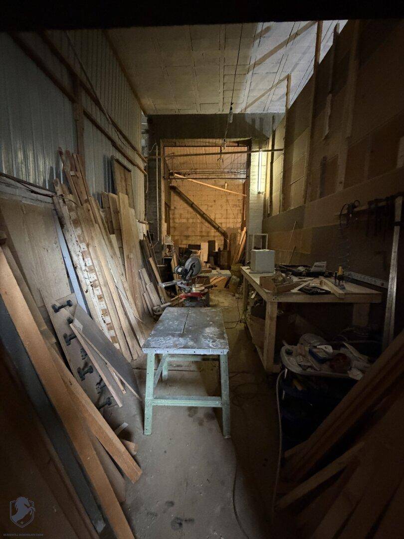 Schmaler Werkstattgang mit Holzbrettern an den Wänden und einer Werkbank mit Säge in der Mitte.