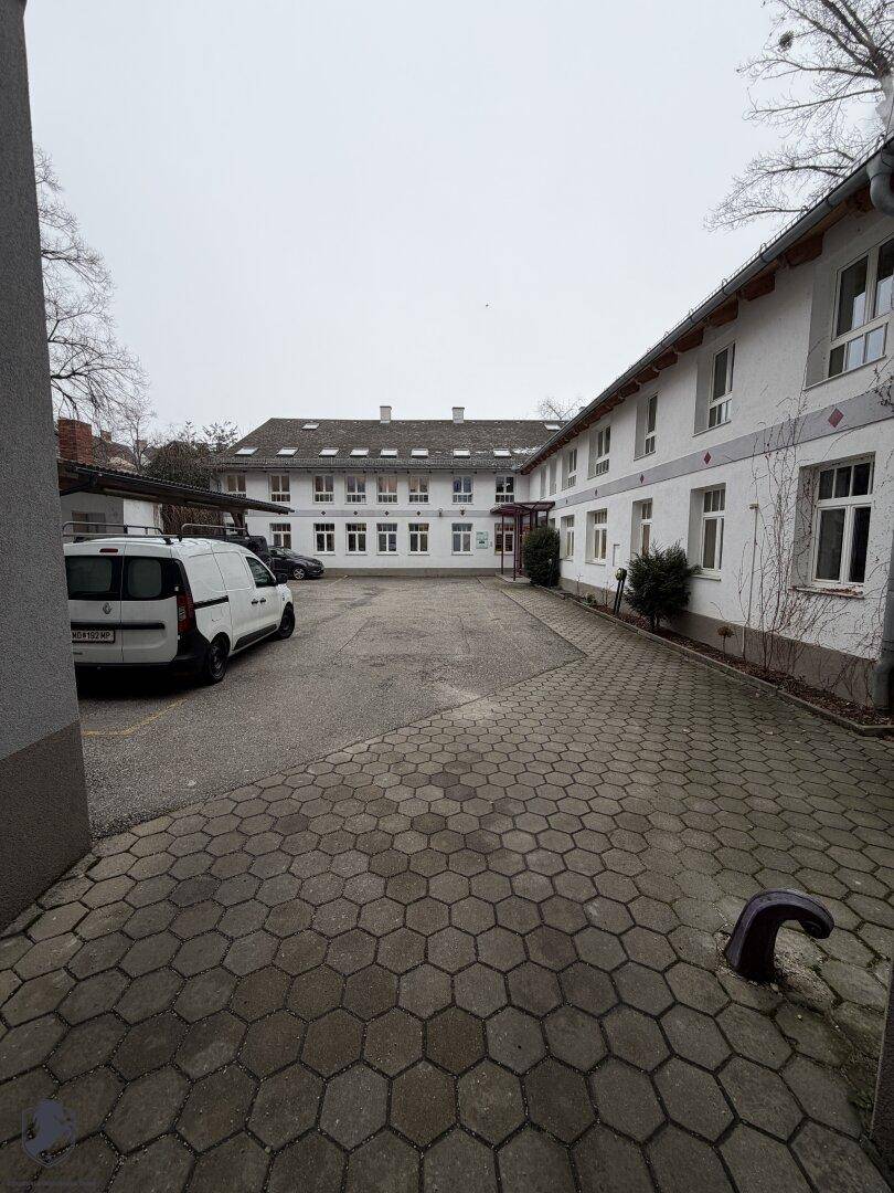 Großer gepflasterter Innenhof mit Parkplätzen und weißem Gebäude im Hintergrund.