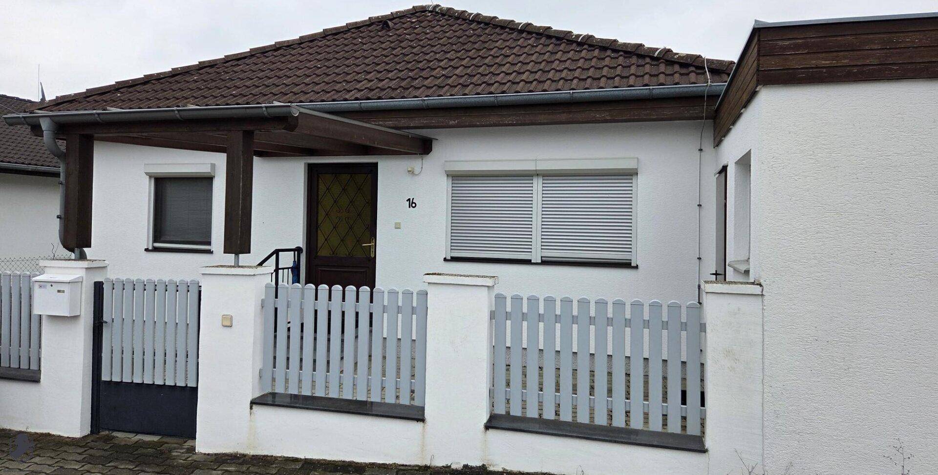 Einfamilienhaus mit weißer Fassade, braunem Ziegeldach und einem kleinen Vorgarten mit Zaun.
