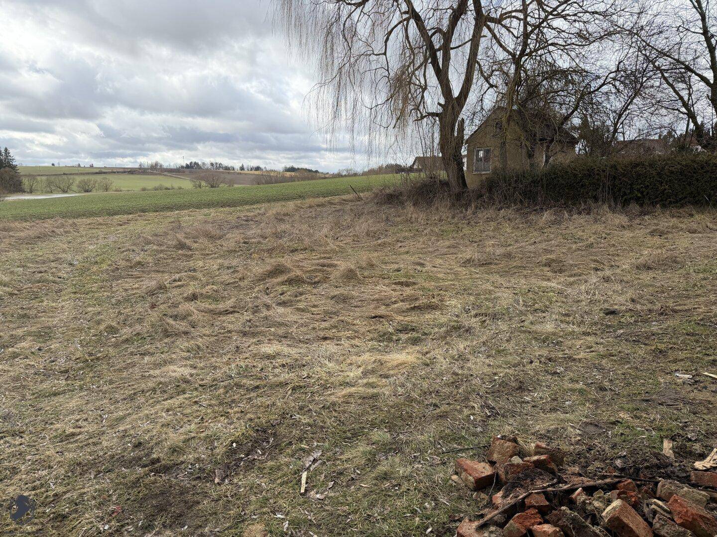 Weitläufiges Feld mit trockenem Gras und einem Haus in der Ferne unter bewölktem Himmel.