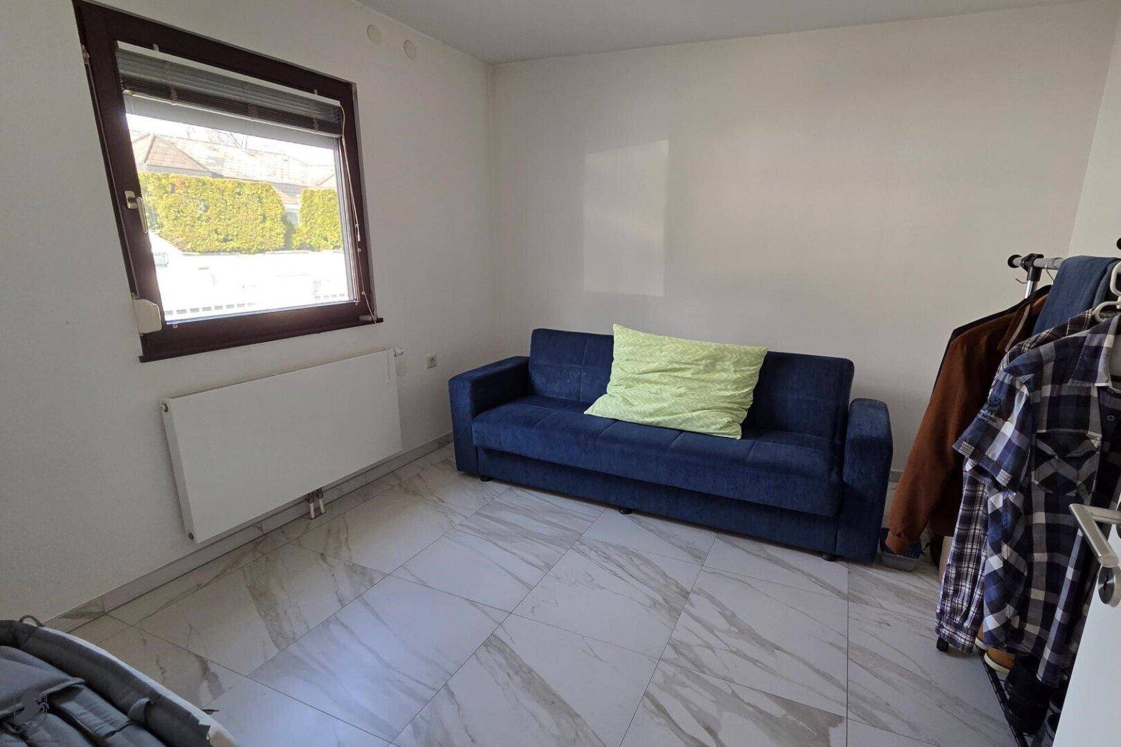 Kleines Zimmer mit blauem Schlafsofa, Fliesenboden und Fenster mit Blick ins Grüne.