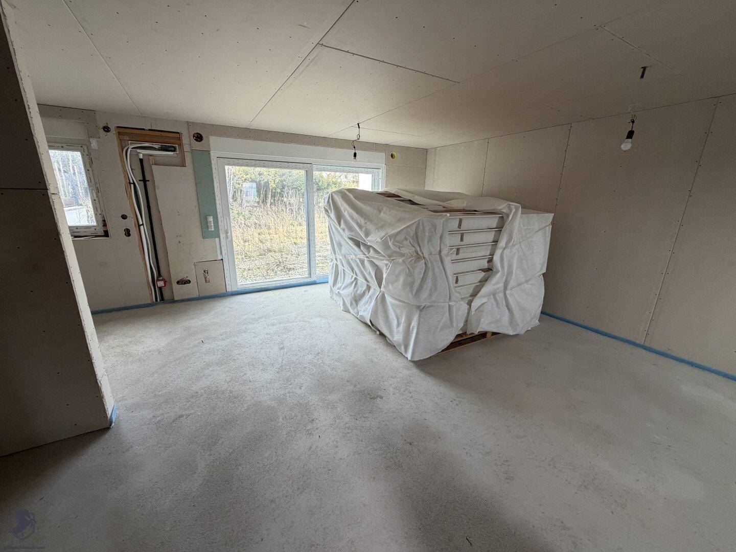 Geräumiger Wohnbereich im Rohbau mit großen Fenstern und Baumaterialien.