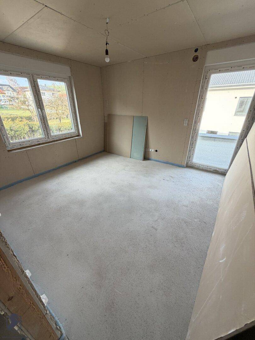 Geräumiges Zimmer im Rohbau mit mehreren Fenstern und Zugang nach draußen.