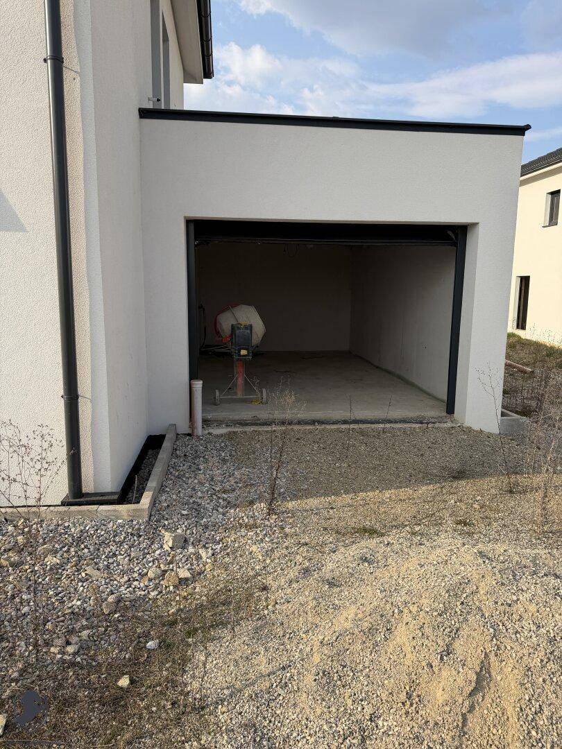 Offene Garage im Rohbau mit Betonboden und Baumaschinen.