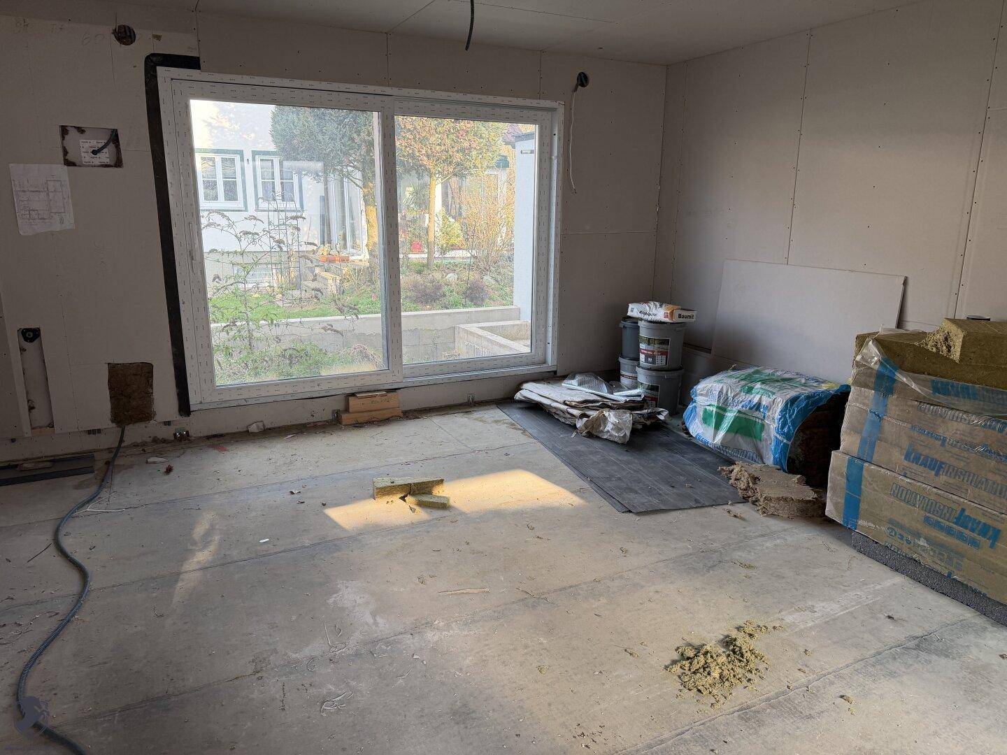 Geräumiger Wohnbereich im Rohbau mit großen Fenstern und Baumaterialien.