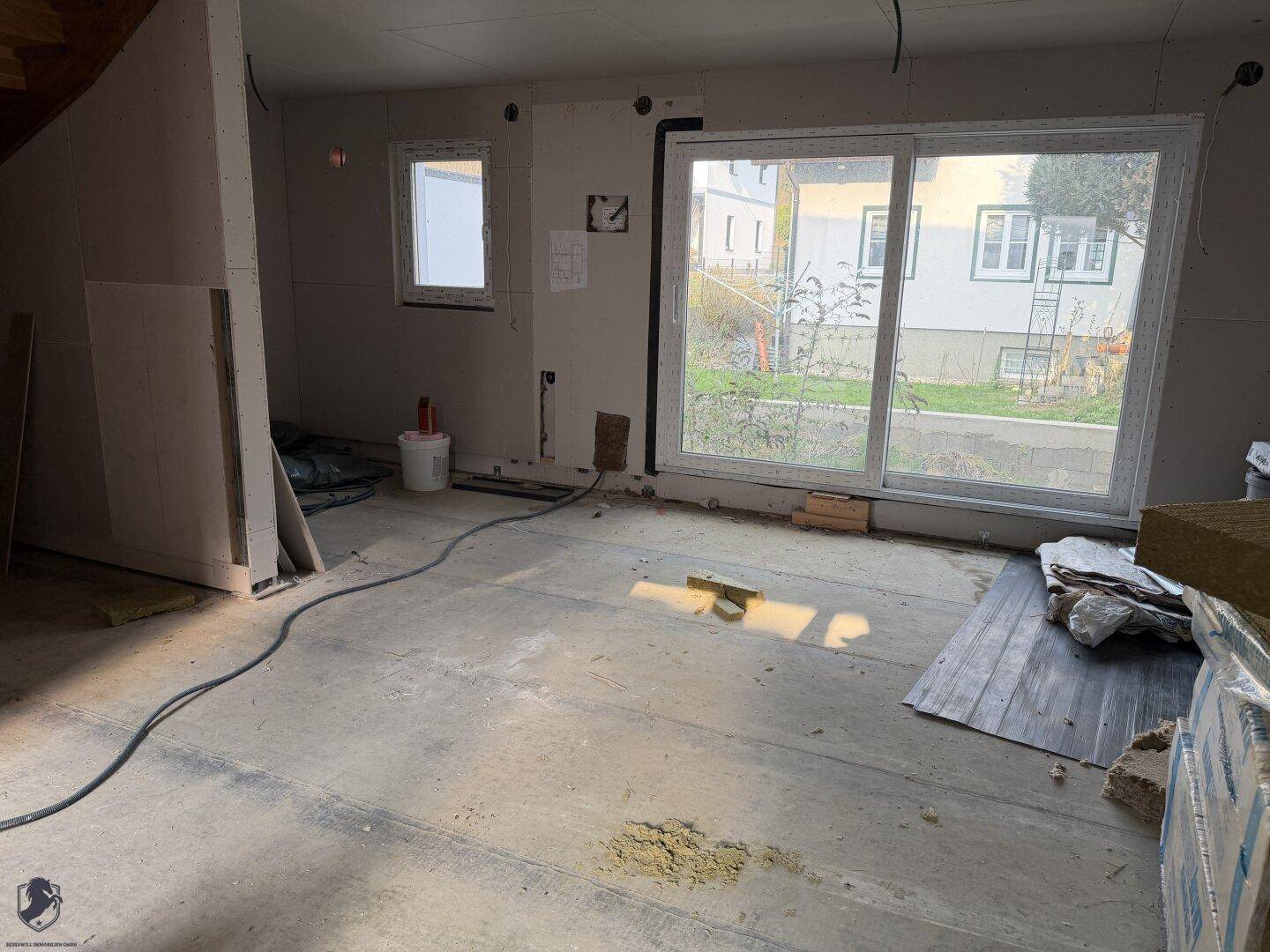 Geräumiger Wohnbereich im Rohbau mit großen Fenstern und Baumaterialien.
