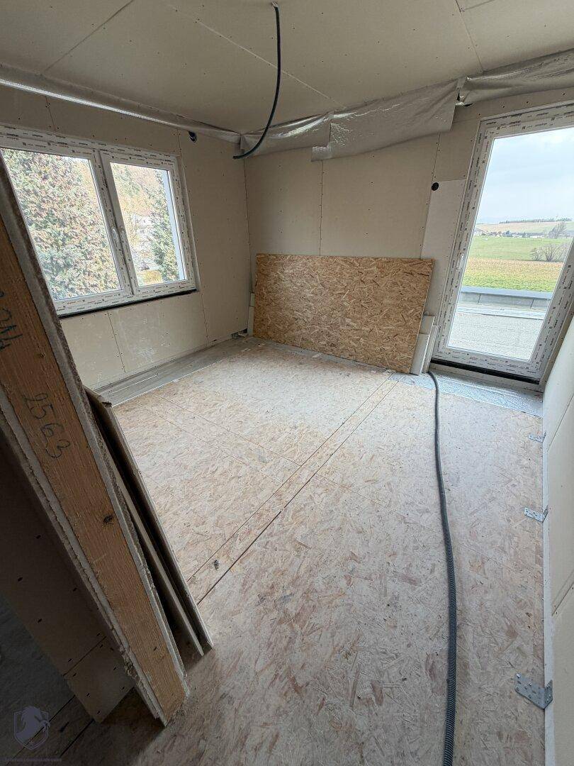 Zimmer im Rohbau mit Fenstern und Zugang zu einem möglichen Dachterrassenbereich.