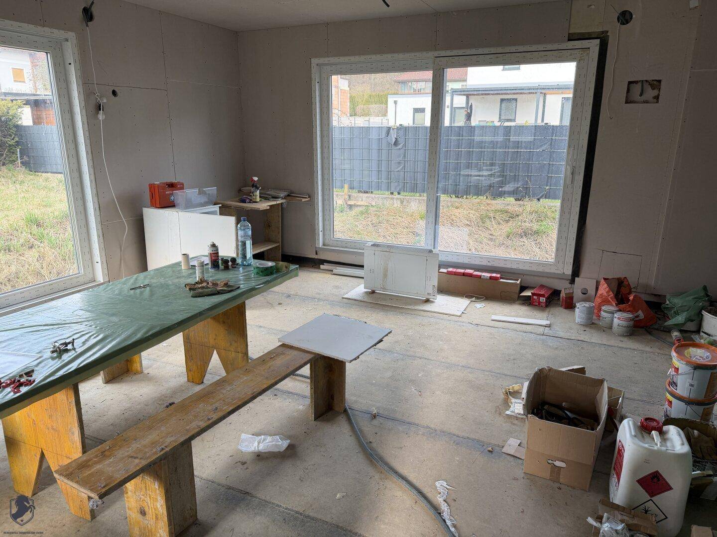 Geräumiger Wohnbereich im Rohbau mit großen Fenstern und Baumaterialien.