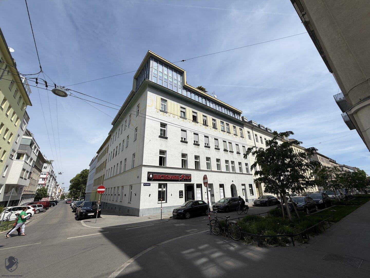 Weitwinkelansicht des Eckgebäudes mit moderner Dachaufstockung und parkenden Autos auf der Straße.