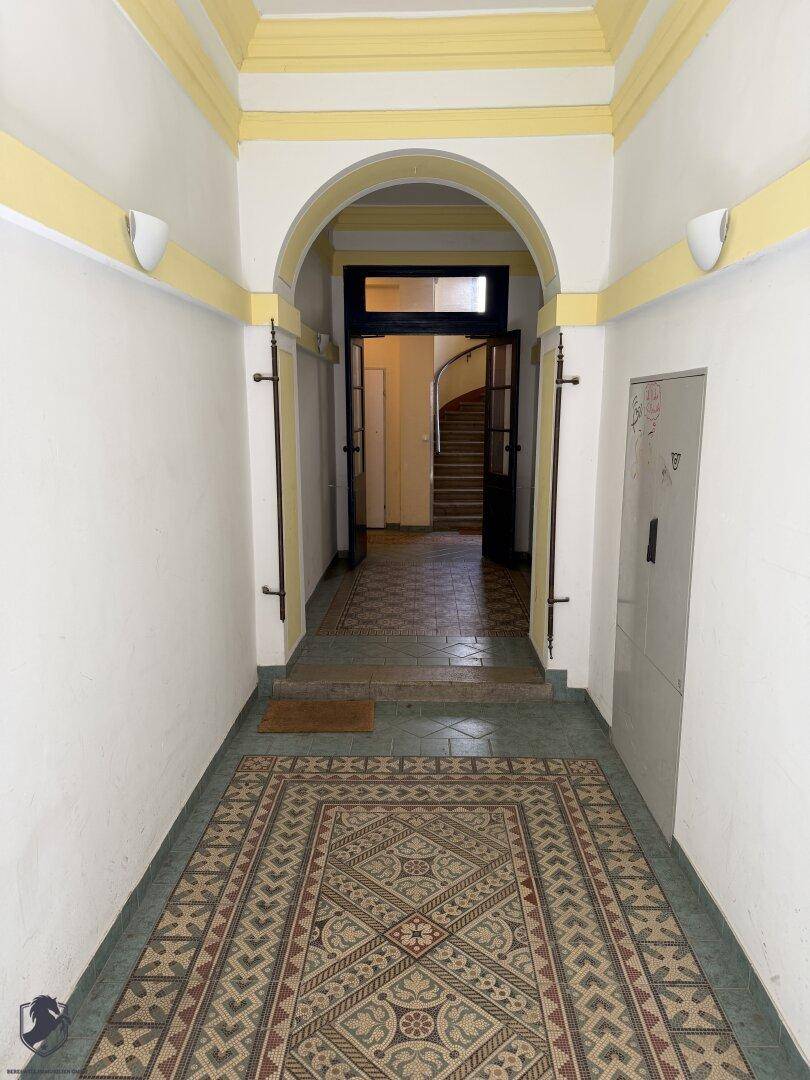 Historischer Eingangsbereich mit gemusterten Fliesen und einem Bogen, der zum Treppenhaus führt.