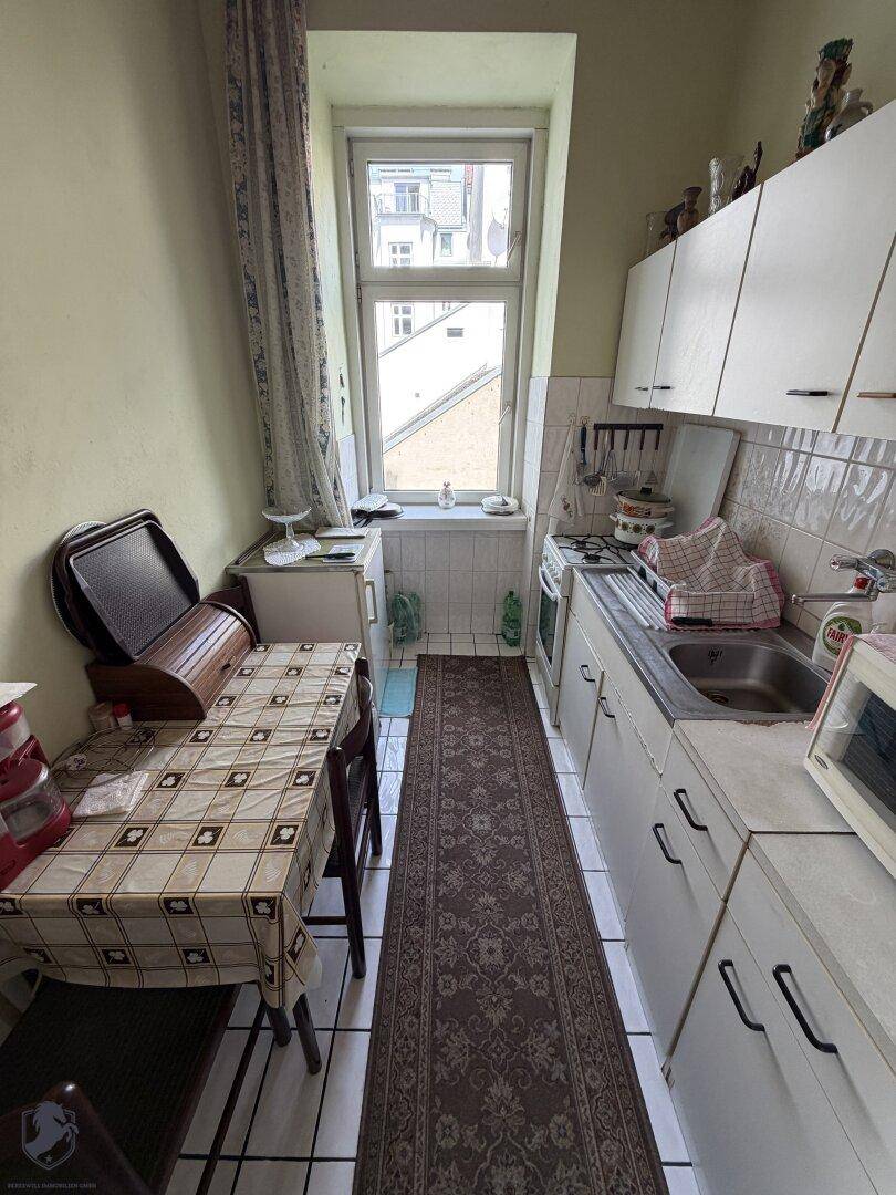 Schmale Küche mit weißen Schränken, Fliesenboden und einem Fenster mit Blick nach draußen.