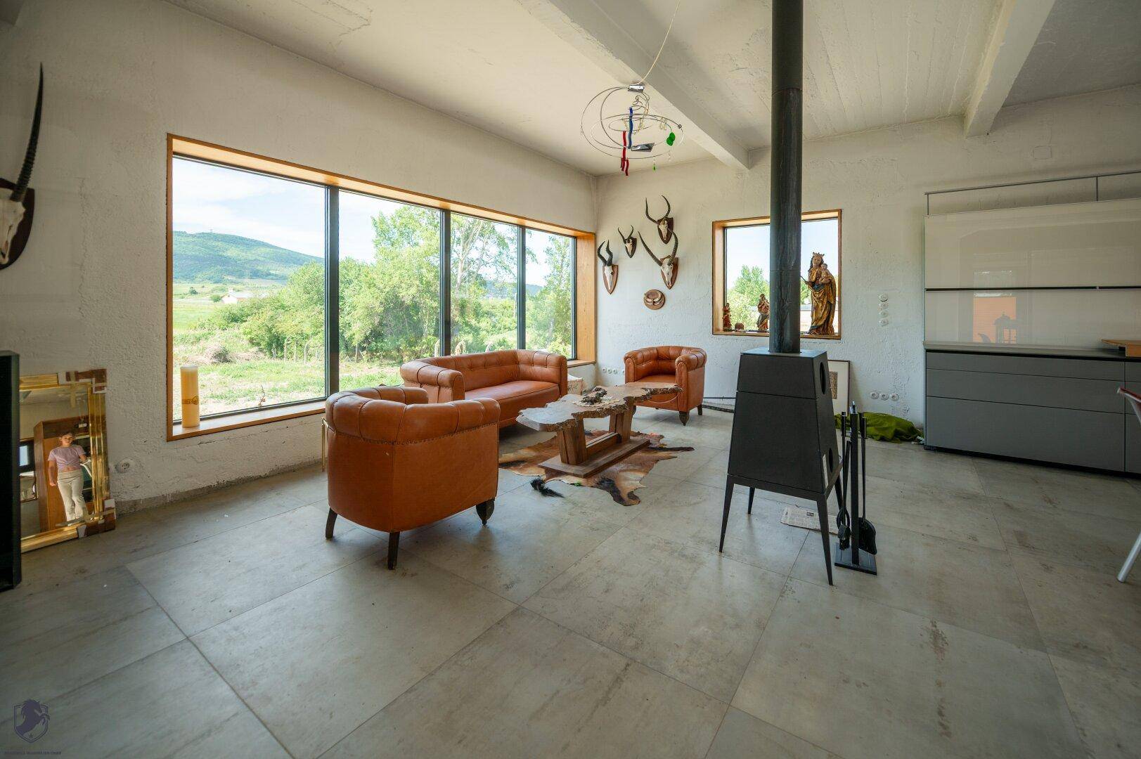 Offener Wohnbereich mit Kaminofen, Ledermöbeln und Blick auf die moderne Küchenzeile sowie die grüne Landschaft.