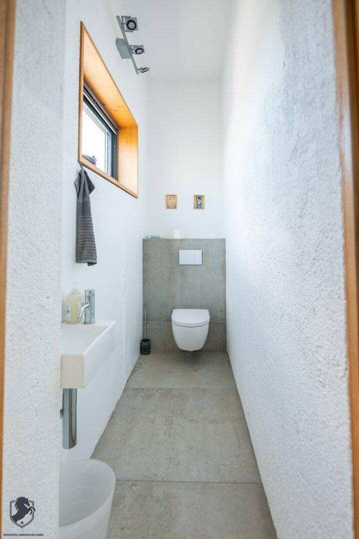Schmale Gästetoilette mit wandhängendem WC, kleinem Waschbecken und einem Fenster für Belüftung.