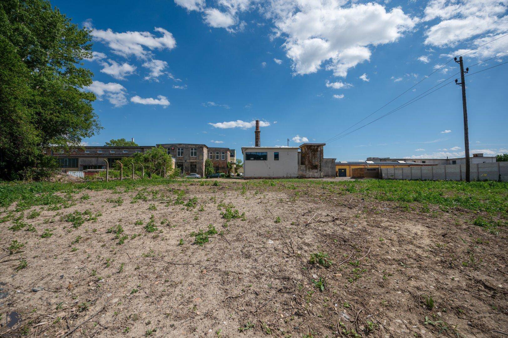 Offenes Gelände mit Gras und Erde, im Hintergrund sind Gebäude und ein hoher Industrieschornstein zu sehen.