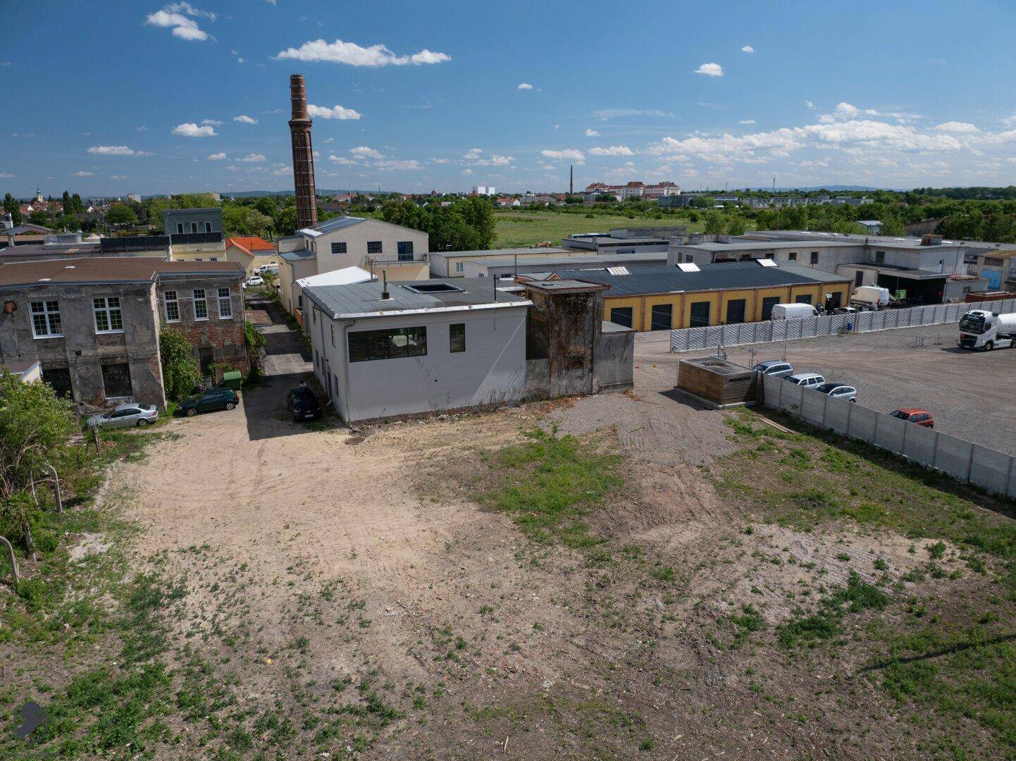 Luftaufnahme eines Industriegebiets mit einer großen unbebauten Fläche und einem hohen Schornstein im Hintergrund.