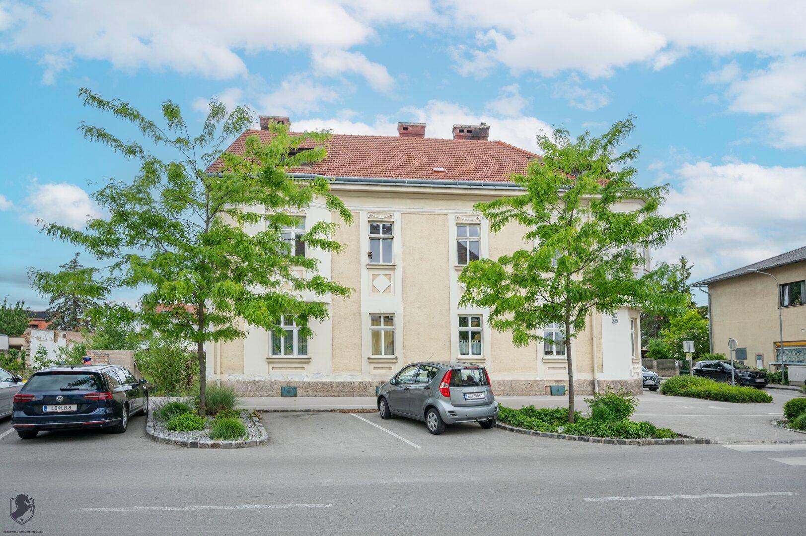Rückansicht des historischen Gebäudes mit Parkplätzen und Bäumen unter einem bewölkten Himmel.