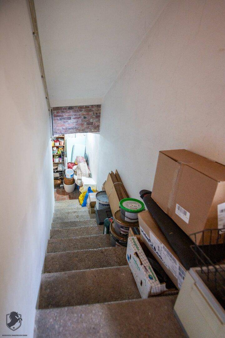 Treppenabgang zu einem Lagerbereich im Keller, umgeben von weißen Wänden und Kartons.