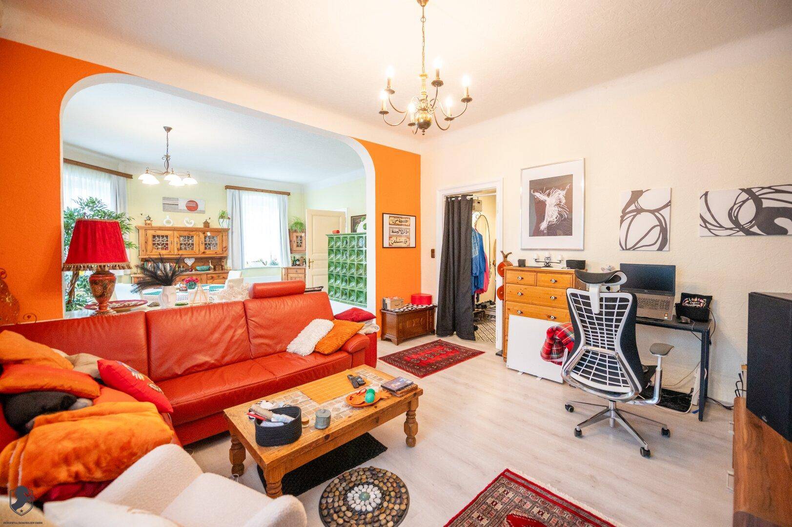 Offener Wohn- und Essbereich mit rotem Sofa, Schreibtisch und Kachelofen.