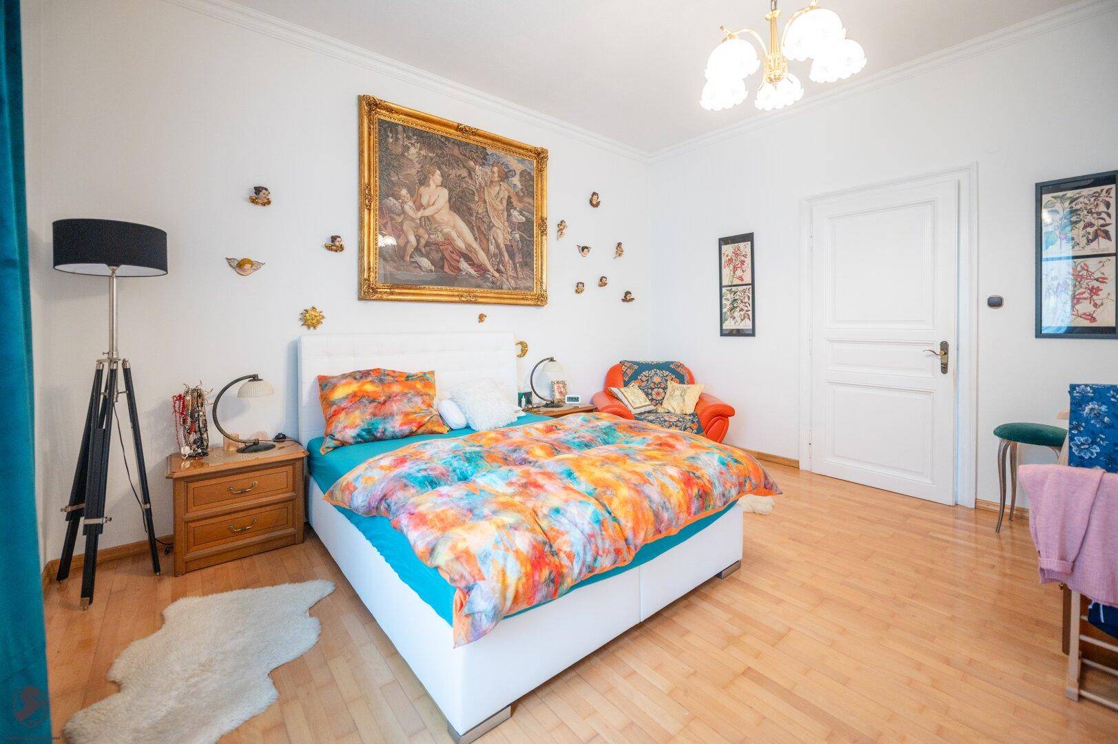 Geräumiges Schlafzimmer mit Holzboden, farbenfroher Bettwäsche und klassischem Kunstwerk.