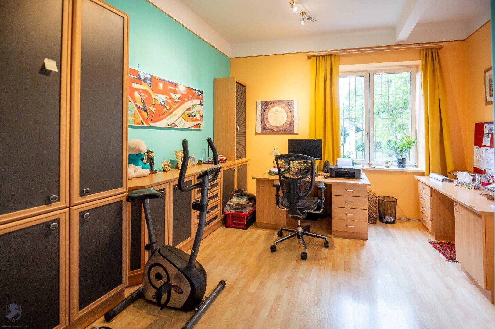 Helles Arbeitszimmer mit Holzboden, Schreibtisch und Fitnessgerät.