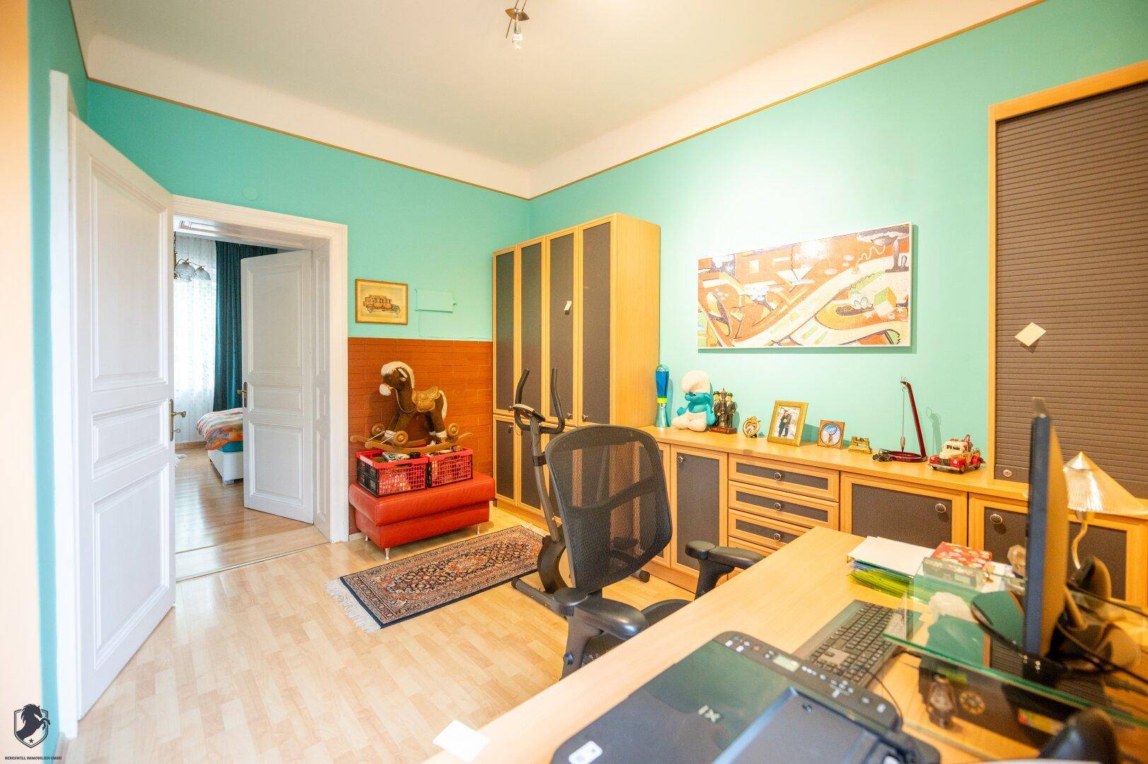 Arbeitszimmer mit Schreibtisch, Regalen und Blick in ein angrenzendes Schlafzimmer.