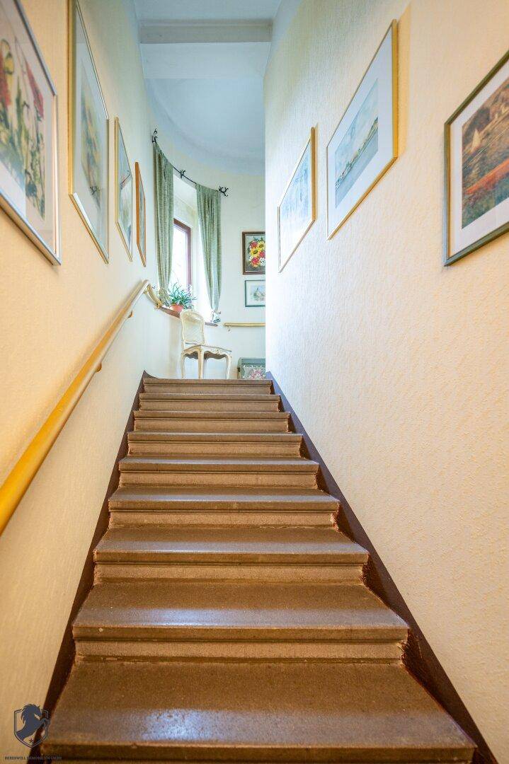 Treppenhaus mit hellem Anstrich, Holzgeländer und Kunstwerken an den Wänden.