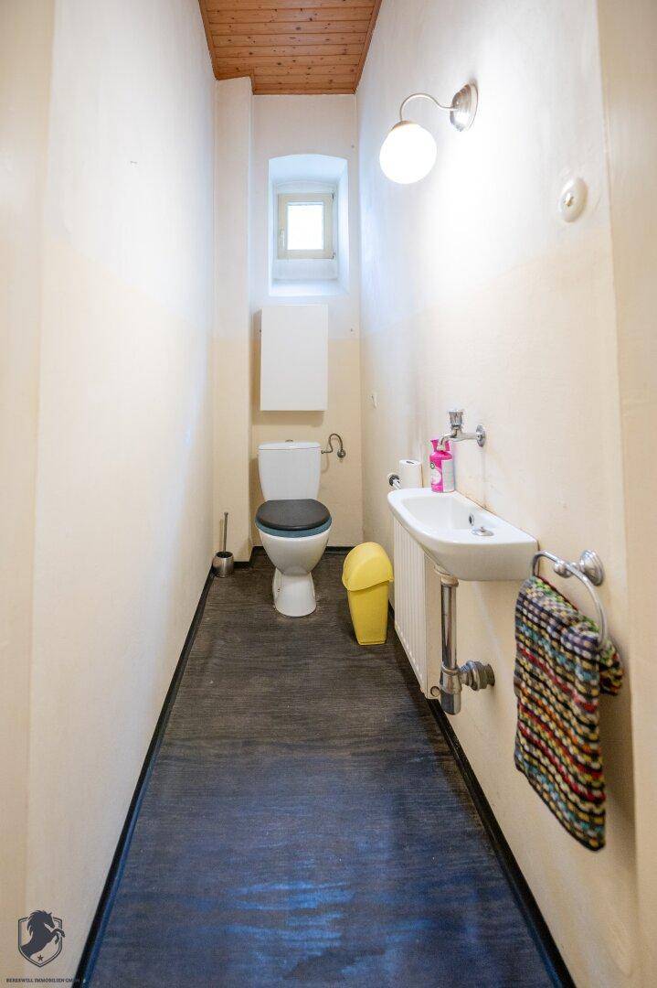 Schmaler Toilettenraum mit Fenster, kleinem Waschbecken und Holzbalkendecke.