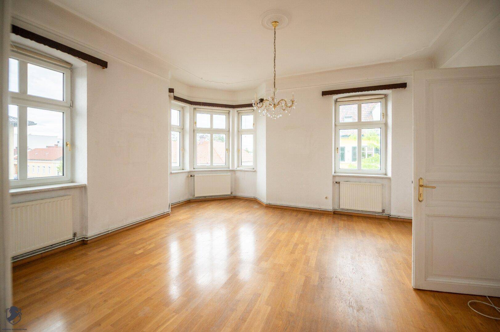 Geräumiges Wohnzimmer mit hellem Parkettboden, großen Fenstern und viel Tageslicht.