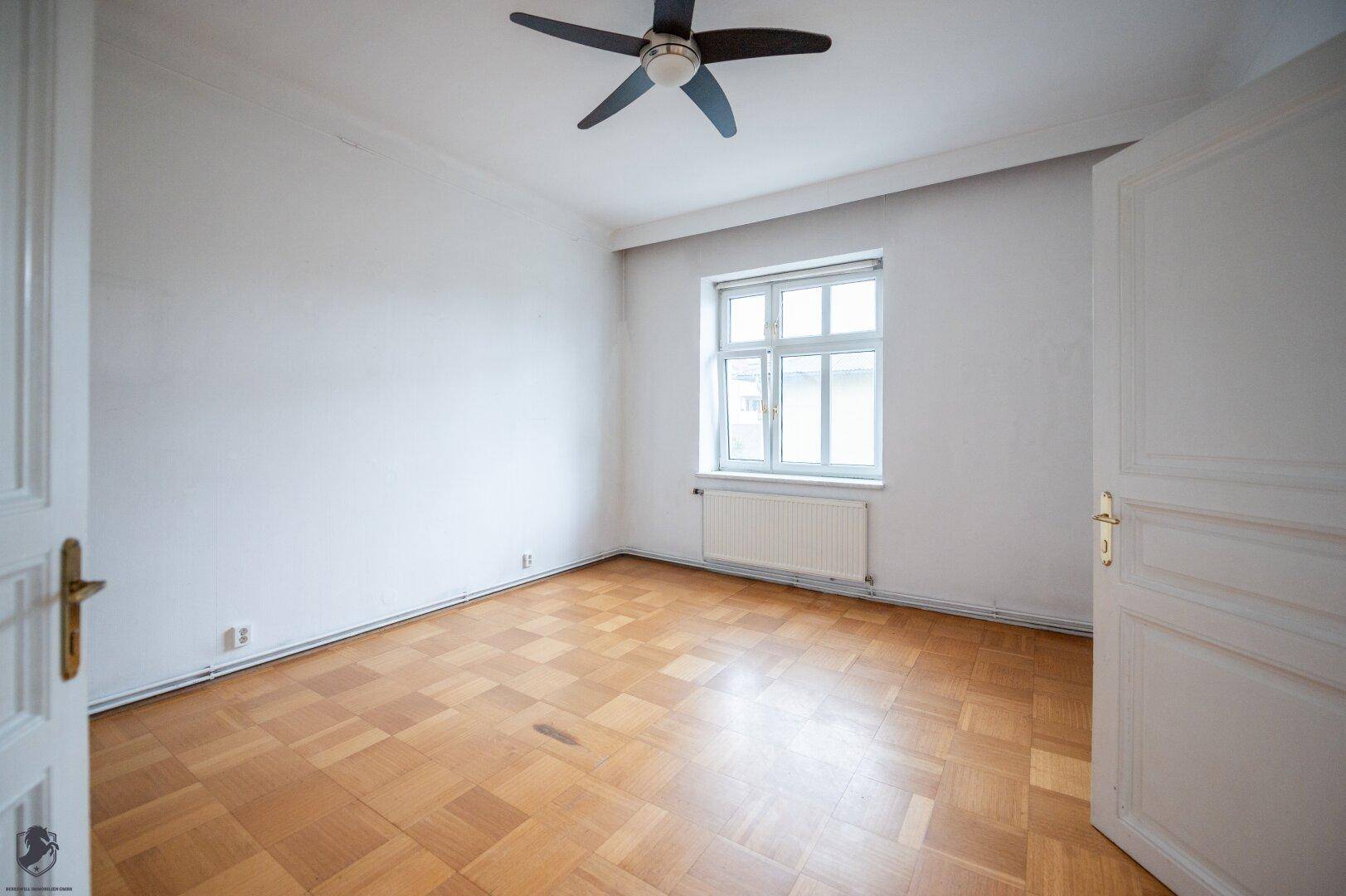 Schlafzimmer mit Parkettboden, Fenster und Deckenventilator, bereit zur Einrichtung.