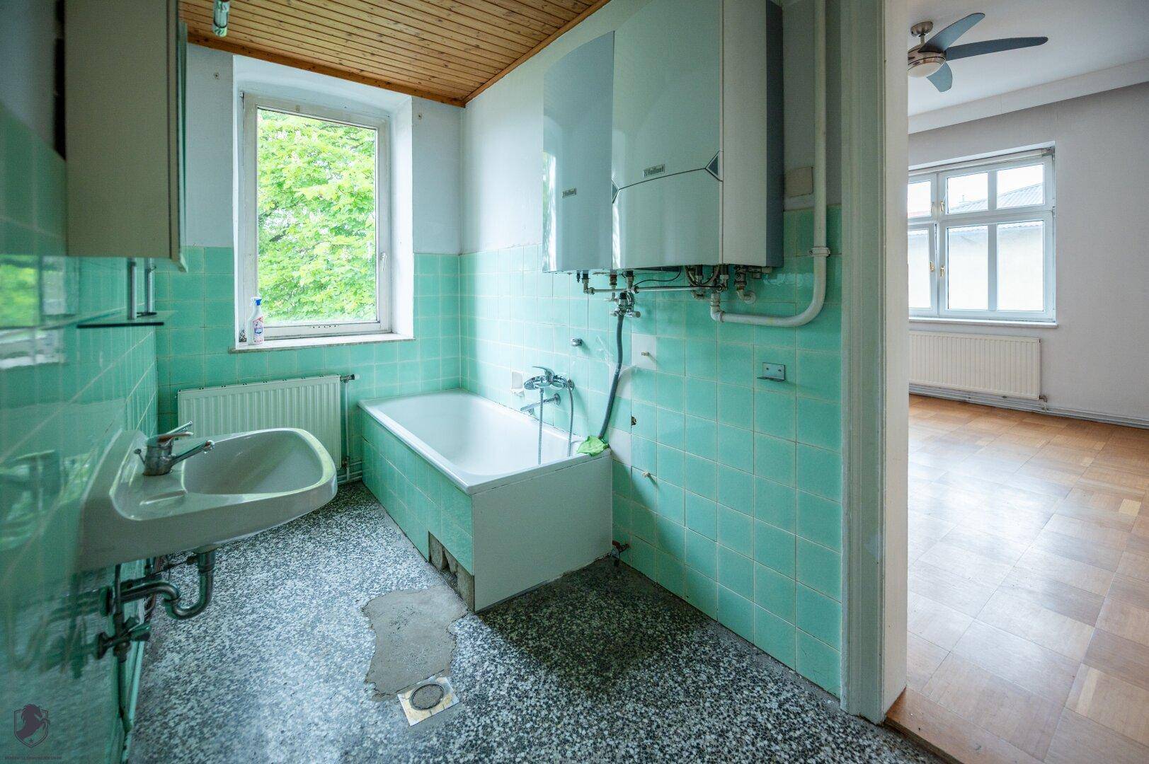 Badezimmer mit mintgrünen Fliesen, Badewanne, Waschbecken und Fenster, renovierungsbedürftig.