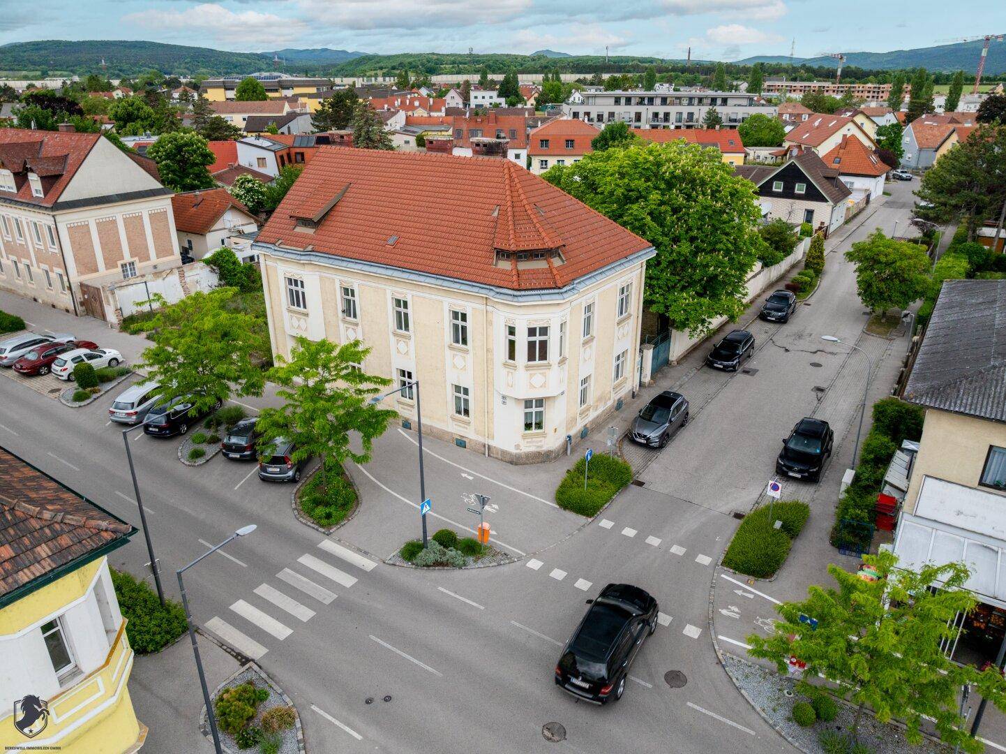 Luftaufnahme eines Eckgebäudes an einer Straßenkreuzung mit umliegenden Häusern und viel Grün.