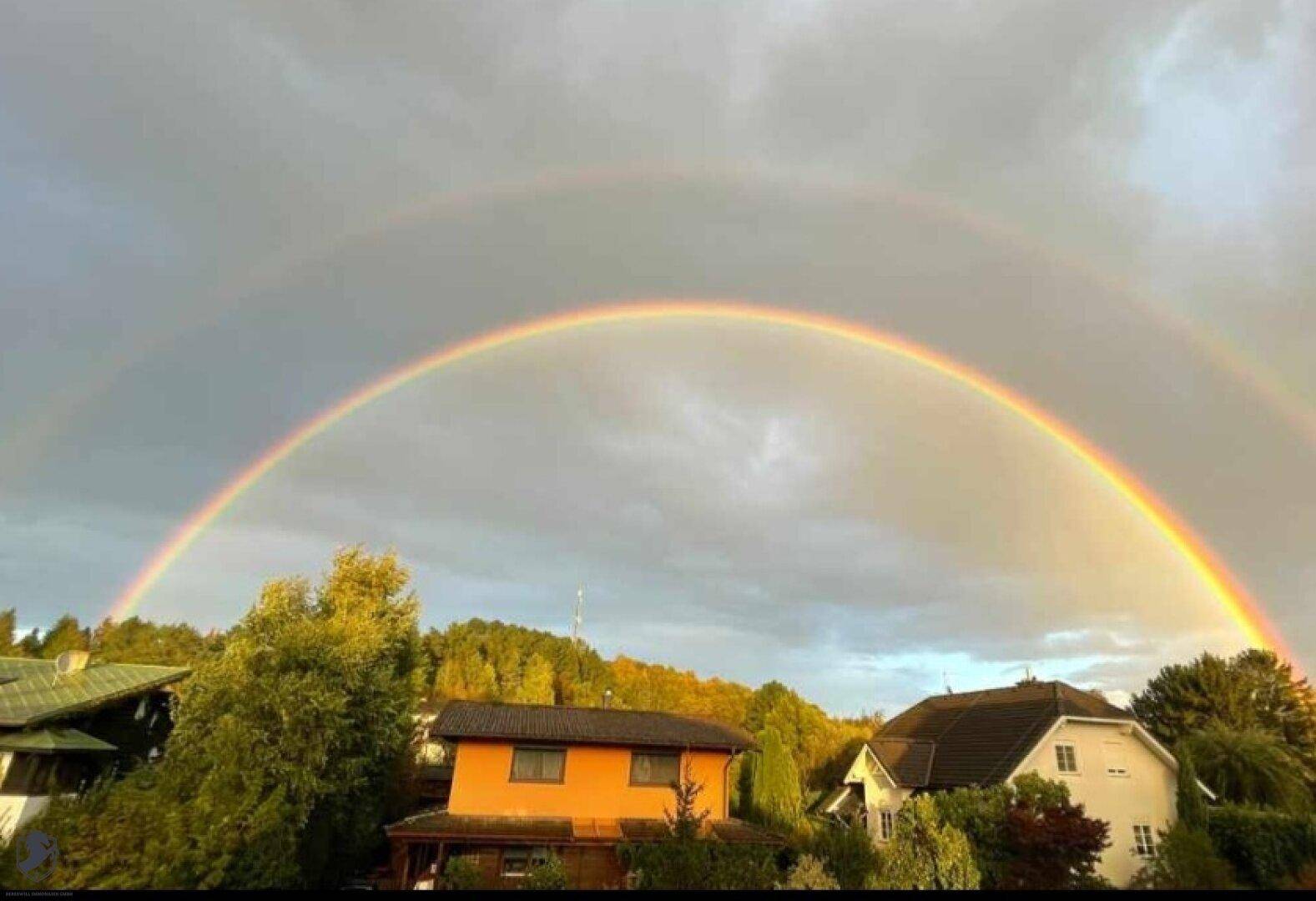 Spektakulärer doppelter Regenbogen über Wohnhäusern und bewaldeten Hügeln bei Sonnenuntergang.