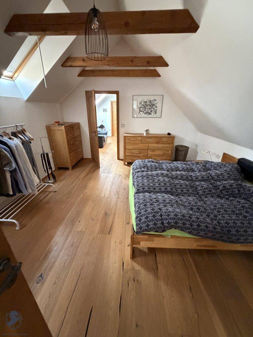 Schlafzimmer mit Holzboden, sichtbaren Holzbalken, Bett und Holzkommode unter einer Dachschräge.