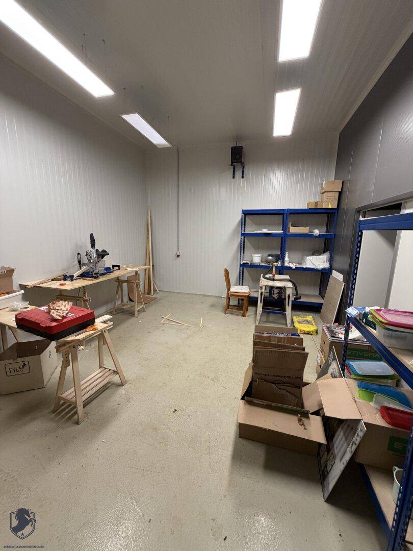 Teil eines Werkstatt- oder Lagerbereichs mit Arbeitstischen, Regalen und Kartons auf Betonboden.