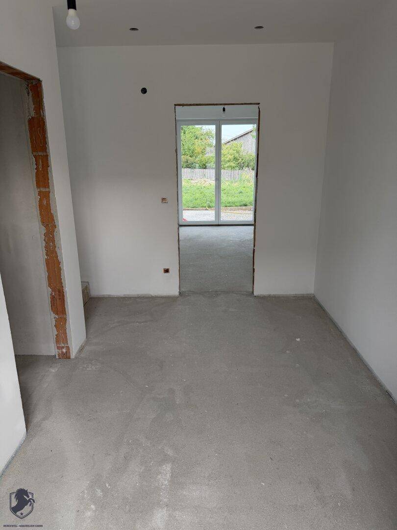 Unfertiger Wohnbereich im Neubau mit Rohbetonboden und Zugang zum Außenbereich.