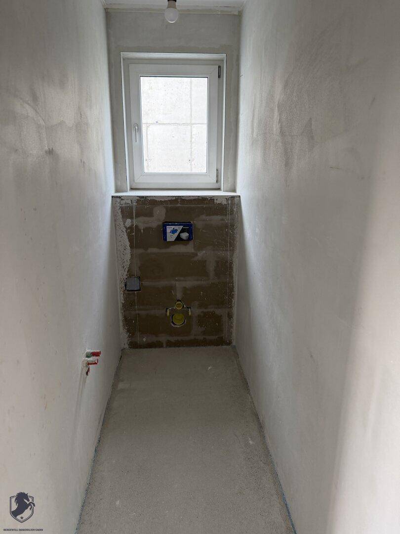 Unfertiger WC-Raum mit Fenster und vorbereiteten Anschlüssen für eine Wandtoilette.