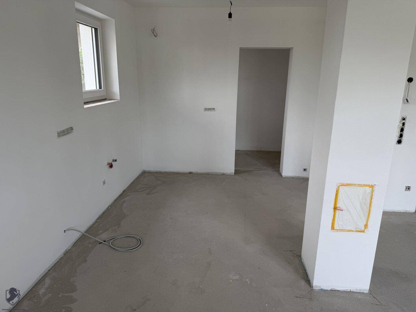 Geräumiger Küchenbereich im Rohbau mit Fenster und vorbereiteten Anschlüssen für die Küchenzeile.