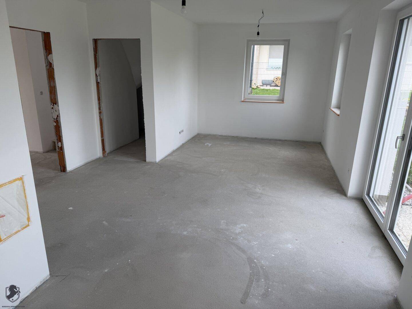 Heller Wohnbereich im Neubau mit Rohbetonboden und großen Fenstern zum Garten.
