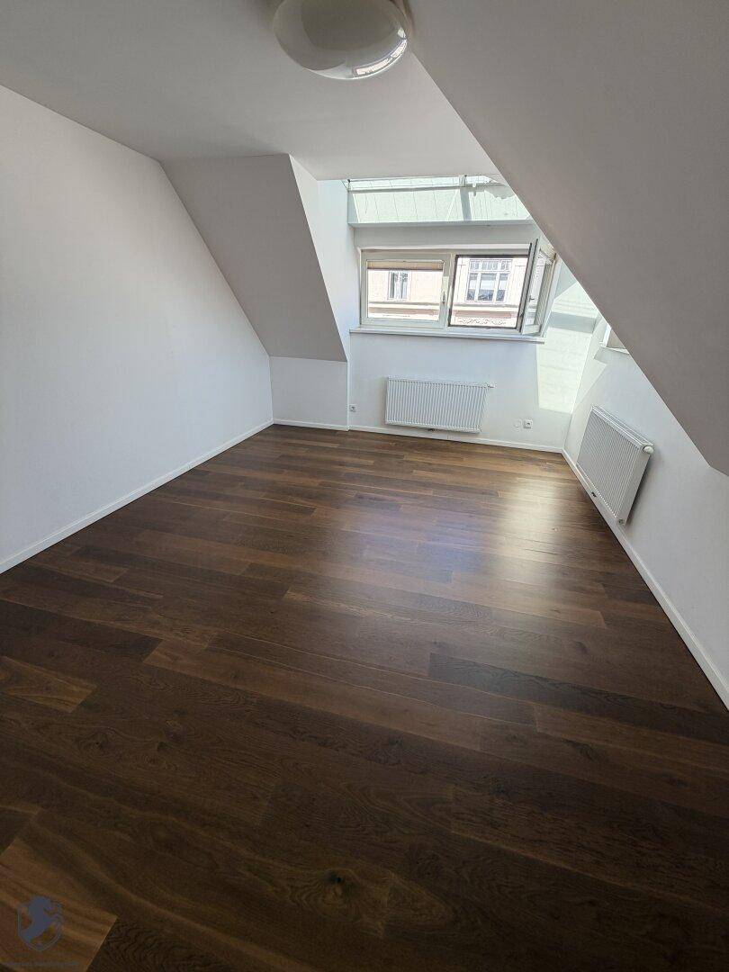 Geräumiges Zimmer im Dachgeschoss mit dunklem Parkettboden und Fenster.